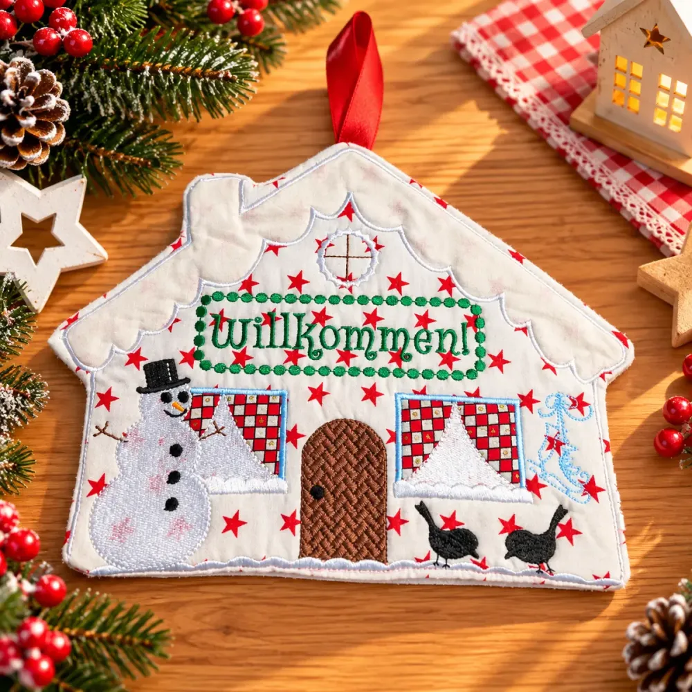 Besticktes Weihnachtshäuschen mit grünem Willkommen-Schriftzug, Schneemann, roten Sternen und zwei schwarzen Vögeln